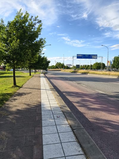 The image depicts a paved sidewalk next to a road in the city of Linköping, Sweden. The sidewalk is made of rectangular pavers, with a section of white pavers bordering the grass. Trees line the left side of the sidewalk, casting shadows on the pavement. The road is a multi-lane asphalt road with a red lane marking. Overhead blue road signs indicate directions to "Ettinge," "E4 34," "Berg," "Tornby N," "Koxviksgatan," "IKEA," and "Ultuna." In the background, there are more trees, some buildings, and flags. The sky is blue with scattered white clouds. The lighting suggests it is daytime, likely late afternoon given the shadows. There are no people visible in the image.