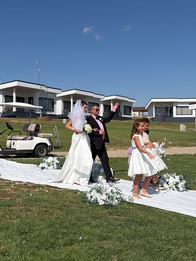 A wedding procession walks down a white aisle runner on a grassy lawn under a clear blue sky. A bride in a white gown and veil is escorted by a man in a black suit and pink shirt. Two young girls in white dresses, presumably flower girls, walk ahead of them, scattering petals. In the background, modern white buildings with dark-framed windows are visible, and a white golf cart is parked to the left of the aisle. The scene is set in Banya, Bulgaria.