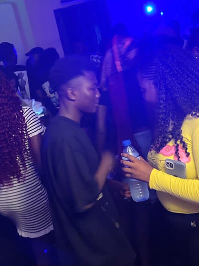 The image depicts a dimly lit indoor scene, possibly a nightclub or party, with predominantly blue and purple lighting. In the foreground, a young man with short, dark hair is seen from the side, wearing a dark t-shirt. To his right, a young woman with long, curly dark hair is visible, wearing a bright yellow long-sleeved top with a cartoonish design and black pants. She is holding a clear plastic bottle of water in her right hand and a light gray smartphone in her left hand, with the phone's camera visible. Another person with long, dark curly hair and a striped t-shirt is partially visible on the left side of the frame. Other individuals are present in the background, largely obscured by the low lighting and the presence of other people. The overall atmosphere suggests a social gathering. The location is identified as Wumat, Nigeria.