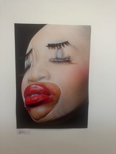 A close-up photograph of a person's face, angled in profile, is displayed on a white wall in what appears to be an art exhibition. The face is heavily made-up, with bright red lipstick on the lips, blue eyeshadow, and exaggerated false eyelashes. The skin texture is overlaid with a fine, ribbed pattern, creating an artificial, mask-like appearance. The background of the photograph is dark, creating a stark contrast with the illuminated face. Below the photograph, a small white label is affixed to the wall, containing text that reads "Muskan Verma BA Makeup and Hair Design." This suggests the artwork is part of a student exhibition. The lighting in the room is even and diffused, typical of gallery lighting. The wall itself is painted a neutral white. The photograph is the sole subject visible on this section of the wall. The location in Epsom and Ewell is not identifiable from the image itself, but such an exhibition could be held at a local college, art center, or community gallery within the borough.