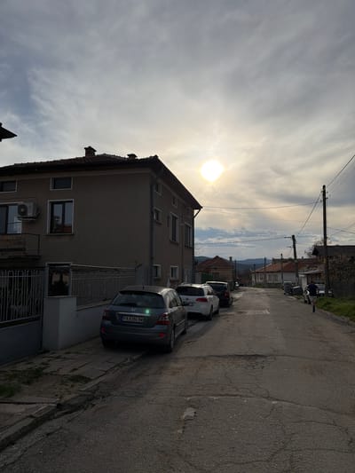 The image depicts a residential street in Popintsi, Bulgaria. A three-story building occupies the left side of the frame, and several parked cars are visible on the right side of the street. The sky is partly cloudy with the sun shining through. Power lines are seen stretching across the top of the image, and distant mountains are visible in the background. The street is asphalt, showing cracks and signs of wear. The front of the building has an air conditioning unit. A pedestrian is walking in the street near the top of the frame. The car closest to the camera has a Bulgarian license plate that reads "PA 8386 BM".
