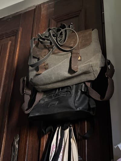 Two bags are hanging on a wooden door. The upper bag is a large, canvas messenger bag with brown leather accents and straps. The lower bag is a black bag, possibly leather or synthetic, with straps. Several other items, appearing to be bags or fabric, are hanging below the two main bags. The door is dark wood with paneling details. This scene is indoors, likely in a residential setting. There is no indication of the specific location within Herceg Novi, Montenegro.