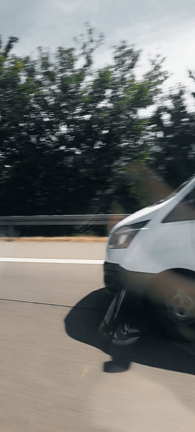 A white van is captured in motion on a highway, its movement evidenced by the blurred trees in the background and the motion blur on the vehicle itself. The van is positioned on the right side of the frame, with its front end and a portion of its side visible. A white line marks the lane on the highway. The sky is overcast. The scene appears to be a typical highway environment, with no specific identifiable landmarks suggesting the city of Endersbach, Germany.
