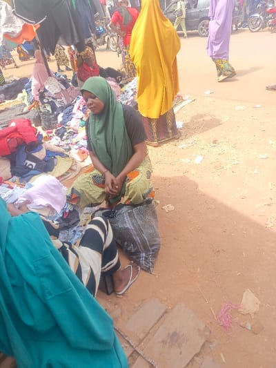 A woman wearing a green hijab and patterned pants sits on the ground in a bustling outdoor market. She has a black shirt and her hands are clasped in her lap. Nearby, a pile of clothes surrounds her. Other people wearing traditional clothing are milling around. A large bag rests near her. The ground is reddish-brown and slightly uneven. Garments hang above, creating a makeshift canopy.