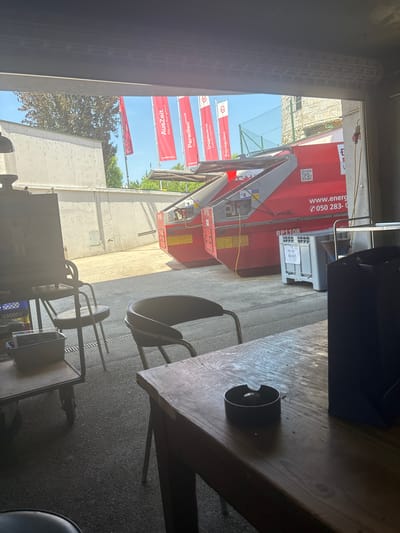 This image depicts an outdoor scene, possibly a loading dock or garage area, in Gebersdorf, Austria. In the foreground, a wooden table with a dark cylindrical object, likely an ashtray, sits in shadow. Behind it, several chairs with metal frames and dark seats are arranged.
The middle ground is dominated by a large, bright red industrial container or trailer with "RP1108" visible. Text on the container includes "www.energ" and a phone number "050 283-0". A QR code is also present. There's a grey plastic bin with text on it, seemingly related to waste disposal.
In the background, several red banners are suspended, displaying text in German. One banner reads "AusZeit Sauna-Bergdorf", another "Paradiso****s Hotel", and a third "Tropicana Cabrio-Therme Aquapuloo". The presence of these banners suggests a connection to a leisure facility or resort.
The setting appears to be a well-lit outdoor area, likely during the day given the bright sunlight and shadows. The overall impression is one of a functional service area adjacent to a recreational establishment. No people are visible in the frame.