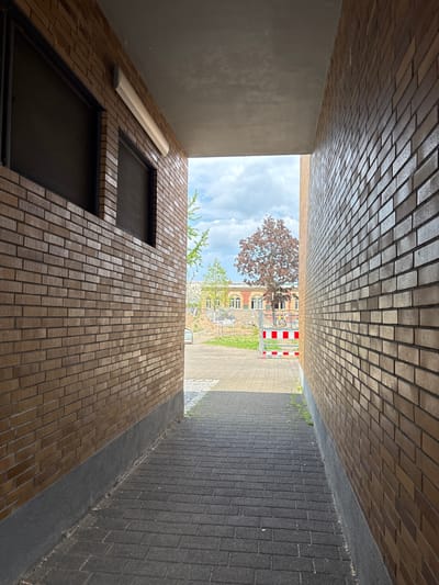 This image depicts a narrow walkway between two brick buildings, leading out to a street scene in Mannheim, Germany. The walkway is paved with dark gray interlocking bricks. The brick walls on either side are light brown with visible mortar lines. The left wall has two dark-paned windows and a cream-colored light fixture running horizontally above them. The ceiling above the walkway is a smooth, light gray concrete.
In the distance, a street or plaza is visible. There are trees, including one with reddish-brown foliage, and a building with arched windows. Construction fencing with red and white striped barriers is present in the middle ground. The sky is partly cloudy with a blue sky visible. There are no people visible in the image. The scene appears to be an urban environment, possibly a side street or alleyway.