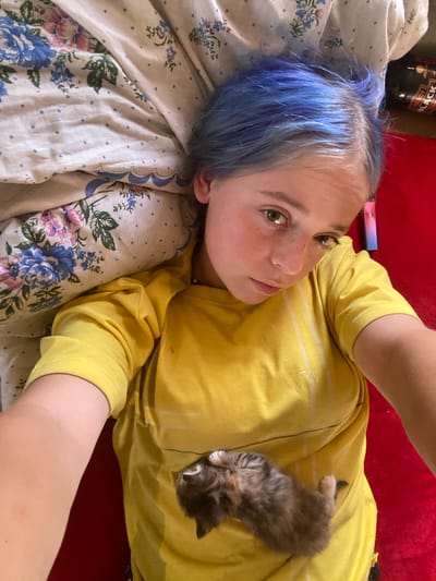 A young person with blue-dyed hair lies on their back, looking up at the camera. They are wearing a yellow t-shirt and are nestled against floral patterned bedding. A tiny kitten is curled up on the person's chest. The person's left arm is resting on the bedding, and their right arm is extended as if taking the selfie. The background is a red surface, possibly a bedspread or blanket. A black energy drink can is visible in the upper right corner. The scene appears to be indoors, in a bedroom. The location in Nova Praha, Ukraine is not discernible from the image.