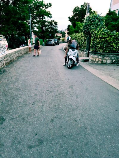 This media file, filmed in Đenovići, Montenegro, depicts a street scene with a focus on people and a scooter. The image is oriented vertically.
In the foreground, a white scooter is prominently featured. A person, wearing a helmet and a light-colored top with dark pants, is seated on the scooter, appearing to be either about to depart or having just arrived. Their posture suggests casualness, and the helmet indicates a concern for safety while riding.
Further in the background, two individuals are standing near a low stone wall adorned with lush greenery. One person, wearing a dark green t-shirt and shorts, stands upright, holding a bag. The other person, in a white top and shorts, is also standing, looking towards the street. Their casual attire and relaxed stances suggest a leisurely moment.
The setting appears to be a street in a residential or coastal area, with buildings visible in the distance. Trees with dense foliage line the left side of the road, creating a natural canopy. The road surface is asphalt, with a clear sidewalk or pedestrian path demarcated by a lighter colored material and a stone border.
The lighting suggests it is daytime, possibly late afternoon or early evening, given the soft illumination and the slightly muted colors. There are no visible signs of distress or urgency; the overall atmosphere seems calm and unhurried. The scene captures a slice of everyday life in a scenic location.