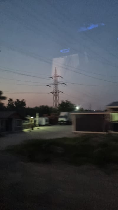 The image depicts a twilight scene with a large electricity pylon dominating the skyline. Below, in the city of Voluntari, Romania, various structures are visible, including what appears to be a small building or shed on the left, a trailer or caravan, and a white truck. In the foreground, there is a dark, blurred area that could be a road or a grassy patch. The overall ambiance is dim, with the sky displaying hues of deep blue and pink, suggesting the transition from day to night. The reflection of blue lights is visible on the upper part of the image, possibly from streetlights or illuminated signs.