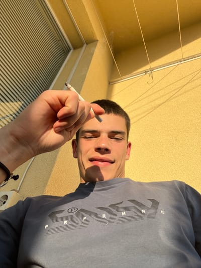 A low-angle, eye-level outdoor shot captures a young man on what appears to be a balcony or terrace in Sopot, Bulgaria. The man, likely in his late teens or early twenties, has short dark hair and a light complexion with visible freckles on his nose and cheeks. He is looking down towards the camera with a relaxed, slight smile. He wears a dark blue/grey long-sleeved shirt featuring a prominent stylized black graphic text across the chest that spells "SUPERMAN" with distinct block letters and vertical line patterns. In his right hand, he holds a lit cigarette, which has a small amount of ash forming at the tip. A black beaded bracelet is visible on his left wrist.
The background consists of a brightly lit, textured yellow wall. Strong sunlight casts long, sharp shadows on the wall, indicating either morning or late afternoon. Below the man, a white horizontal blind or louvered surface is visible, suggesting a window or door. To the left, a clothesline system with horizontal lines and a vertical pole with a hook is present, further confirming the balcony setting. A white two-pin European-style power outlet is partially visible on the lower right of the frame. The overall impression is one of a sunny day.