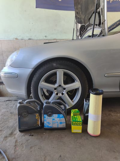 A silver Mercedes-Benz car, with its hood open, is parked in a garage in Svilengrad, Bulgaria. Two black jugs of Mercedes-Benz engine oil, two yellow boxes of Mann Filter engine air filters, and one tall, cylindrical air filter are placed on the concrete floor in front of the car's front passenger-side wheel. The garage has light green walls, and the floor is dusty. The overall scene suggests preparation for car maintenance.
