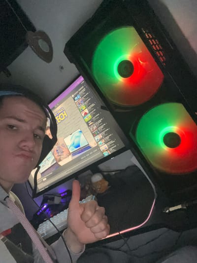 A selfie photograph shows a person with short hair wearing headphones, positioned in front of a computer setup. The monitor displays a screen with various icons and images, including the words "TECH DETAILS" visible at the bottom. Above the monitor are two computer fans with red and green lighting. To the right of the monitor, a keyboard and a hand giving a thumbs-up gesture are visible. The overall environment appears to be an indoor, possibly home-based, setting. The image was taken in Benningen, Germany.