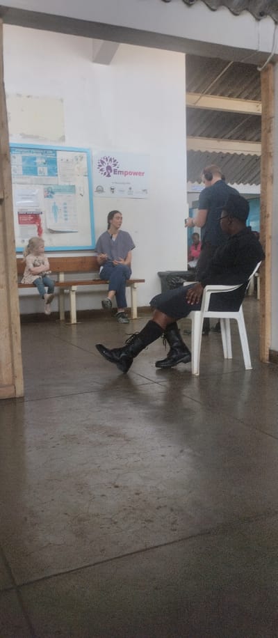 The image shows a scene inside a building in Kisumu, Kenya, located at coordinates -0.09 latitude and 34.771 longitude.  There are four people present, all adults.  The scene shows a woman sitting on a bench, with a young girl seated on the bench next to her.  An adult man is sitting in a white chair to the right of the woman on the bench.  He is wearing a black shirt and black pants.  A second adult man is standing behind the woman on the bench, holding a phone to his ear.  He is wearing a blue shirt and black pants. The interior walls are white, and there are several posters on the wall.  There is a wooden bench and a white chair. The floor is made of concrete tiles.  The scene appears to be inside a waiting room or a similar area of a medical facility or other service institution.