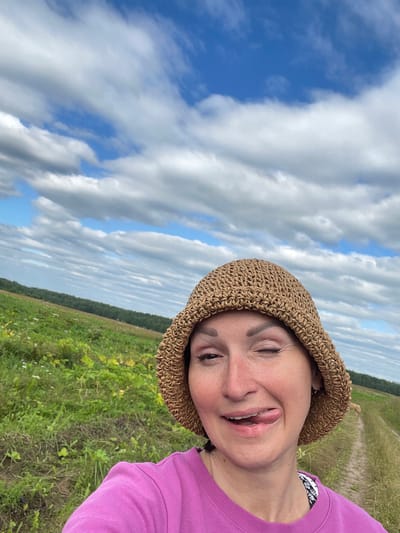 A woman in a straw hat and a pink sweatshirt winks and sticks her tongue out at the camera while standing in a grassy field under a cloudy blue sky. A dirt path runs alongside the field, and a forest is visible in the distance. There is a dog in the background. The image is a selfie taken outdoors, likely in Dresvyanovo, Russia.