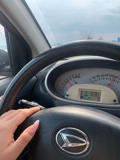 The media file shows a close-up view from inside a car, focusing on the steering wheel and dashboard. A person's hand with long, light-colored nails is visible on the right side, resting on the steering wheel. The dashboard displays a speedometer and a digital screen. The speedometer has markings up to 120, with a red needle pointing between 40 and 60. The digital screen shows "5:38" and "3.8 L/100km," indicating the time and fuel consumption. The exterior view through the windshield suggests it is daytime, with a bright sky and possibly trees in the distance. The overall scene is set within a vehicle, likely in a driving or parked situation, with no strong indication of emotion. The location context provided is Malo Konare, Bulgaria, but this is not directly discernible from the visual elements in the image itself.