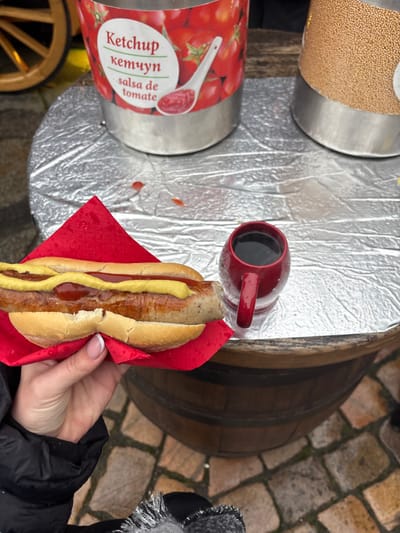 A close-up, first-person perspective shot details a food and beverage transaction at an outdoor stall in Dresden, Germany. A person's right hand, featuring French-tipped manicured nails and wearing a black jacket with a fur-trimmed cuff, holds a prepared hot dog. The hot dog, a grilled sausage nestled in a bun and wrapped in a red paper napkin, is generously topped with parallel lines of yellow mustard and a zig-zag of red ketchup.
To the right of the hot dog, a small, dark red ceramic mug with a handle contains a dark liquid. The food and mug rest on a table surface covered with crinkled silver foil, which bears scattered red liquid drops, likely ketchup. At the back of this surface, two large, cylindrical stainless steel condiment dispensers are visible. The left dispenser is labeled "Ketchup" in English, Russian (кетчуп), and Spanish (salsa de tomate), adorned with tomato imagery. The right dispenser contains a granular, light brown substance, possibly mustard seeds.
The table itself is constructed from a wooden barrel. Below the barrel, the ground is composed of wet cobblestones, indicating an outdoor setting. A portion of a wooden wheel structure is visible in the upper left background.