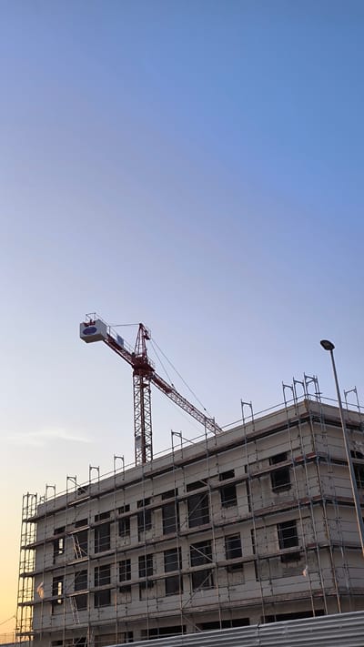 A multi-story building under construction in Ripollet, Spain, is depicted from a low-angle perspective. The building's exterior is enveloped in a comprehensive system of metal scaffolding. A prominent red and white tower crane, branded "COMODA," rises behind the structure, with its jib extending horizontally to the left. The sky overhead transitions from a light, warm blue at the lower left horizon, suggesting either dawn or dusk, to a deeper blue in the upper portion of the frame. A black street lamp with a modern fixture stands on the right, its pole extending above the building's roofline. A corrugated metallic fence or barrier partially obscures the immediate foreground at the bottom of the image. No individuals are visible within the scene.