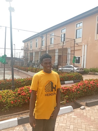 A young man stands outdoors, smiling brightly, under a clear, bright sky on a sunny day. He has dark skin and short, dark curly hair, and is wearing a mustard yellow short-sleeved t-shirt with a black graphic design. The graphic features a stylized "H" composed of horizontal lines, with the word "HENSON" printed in black text below it. He is also wearing dark-colored pants, with his left hand visibly in his pocket.
The man is positioned slightly to the left of the center, standing on a light-colored paved surface with a brick-like pattern, next to a curb painted white and black. Behind him, a low hedge of green bushes with numerous small red flowers stretches across the mid-ground. A chain-link fence runs behind these bushes, separating the foreground from a large, multi-story building in the background.
The building is painted a light peach or orange color and features multiple rectangular windows, some of which have visible grates or security bars. A light-colored car is partially visible behind the bushes on the right side of the image, near the building. On the far left, a tall, dark-colored pole, possibly a light fixture, extends upwards, and some green fabric or plastic is draped over a lower structure nearby. The overall setting appears to be a well-maintained outdoor area, potentially within or near the Akpaka Forest Reserve, Nigeria, as suggested by the location context.