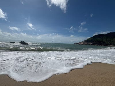 The image captures a coastal scene in Baan Thong Nang, Thailand. The foreground consists of a sandy beach with white foamy waves washing ashore. The water transitions from the foamy nearshore to a deeper blue-green further out. In the middle distance, there are rock formations in the sea and a landmass covered in green vegetation on the right side of the frame. The sky is blue with scattered white clouds. There are no people visible in the frame.
