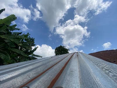 The image captures a low-angle perspective looking upwards from a weathered corrugated metal roof towards a bright blue sky. The prominent foreground features a silvery-grey corrugated roof, likely galvanized iron, showing distinct parallel ridges and valleys. Several reddish-brown streaks of rust are visible along the ridges, suggesting age and exposure.

To the left, large, lush green leaves, characteristic of banana or plantain trees, frame a portion of the sky. Further into the background, other green tree canopies are visible beneath the horizon. On the far right, a section of another roof, darker brown and also corrugated, extends upwards, possibly indicating a different material or more severe weathering.

The sky is clear and a vibrant blue, adorned with scattered white cumulus clouds that appear fluffy and well-defined, reflecting bright sunlight. The strong illumination suggests it is daytime, likely midday or early afternoon. No people or specific activities are depicted. There is no visible text in the image. This scene is indicative of a tropical or subtropical environment, consistent with Achara, Nigeria.