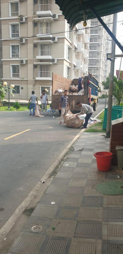 In Chor Sahapur, Bangladesh, several individuals are engaged in collecting and loading refuse into the back of a large truck. The scene unfolds on a paved road adjacent to a sidewalk lined with a green dumpster and a red bucket. The background features a multi-story residential building with numerous windows and air conditioning units. One person, wearing a light blue checkered shirt and patterned trousers, is on the phone while standing near a pile of bags. Another individual, in a blue and white patterned shirt and light-colored trousers, stands beside a large trash bag, preparing to move it. A third person, wearing a gray t-shirt and blue jeans, is also handling a large bag of refuse. Inside the truck, another person is visible, positioned to receive or arrange the waste. Scattered plastic bottles and smaller pieces of trash are visible on the road and sidewalk. The lighting suggests it is daytime.