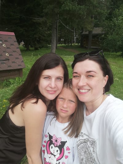 Three females are posing for a selfie outdoors. The person on the left has long dark hair and is wearing a black camisole. The person in the middle is a young girl with blonde hair and is wearing a white t-shirt with a cartoon character on it. She has glitter on her cheeks. The person on the right has dark hair and is wearing a white t-shirt. She has sunglasses perched on top of her head. They are in front of a grassy area with trees and a small wooden structure in the background. The overall setting appears to be a park or recreational area.