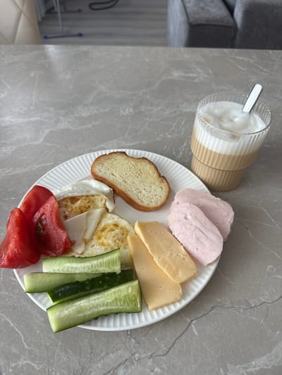 A close-up, eye-level shot captures a breakfast spread on a gray marble countertop. The plate, made of white ceramic with a ribbed texture, holds a variety of food items. In the upper left quadrant of the plate are sliced red tomatoes and two fried eggs, one of which appears to have a slightly runny yolk. To the right of the eggs is a slice of toasted bread. The bottom half of the plate displays sliced cucumbers and several slices of yellow cheese. In the upper right portion of the image, a tall, fluted glass contains a layered beverage, likely coffee with milk or a latte, topped with foam. A silver spoon rests in the drink. In the blurred background, furniture, possibly couches, and a tiled floor are visible, suggesting an indoor setting, likely a home. The image does not contain any information indicating its location in Murom, Russia.