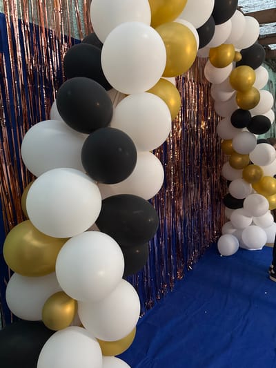 The image depicts an outdoor celebration setup in Sankt Wendel, Germany. A balloon arch, composed of white, black, and gold balloons, is the central focus. The arch extends from the left side of the frame towards the right, partially obscuring a backdrop of shimmering rose gold and blue streamers. The balloons are arranged in a dense, organic formation, creating a festive atmosphere. The ground beneath the arch is covered with a dark blue fabric, possibly a tablecloth or carpet. In the far right of the image, a person's legs, clad in dark pants and shoes with white accents, are visible, suggesting their presence at the event. The overall scene indicates a party or event being held outdoors.