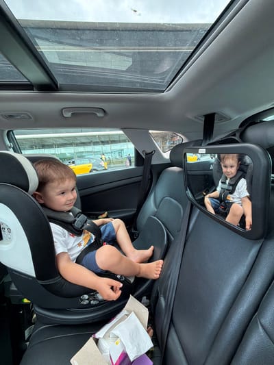 A young boy, approximately three years old, is seated in a black and white child car seat in the back of a vehicle. He is wearing a white t-shirt with a cartoon print and blue shorts. The boy is looking to his left, with his right arm resting on his lap. A black car seat mirror is mounted on the headrest of the seat in front of him, reflecting an image of the boy. The interior of the car features black leather seats and a panoramic sunroof. Outside the car, visible through the windows and sunroof, is an overcast sky and what appears to be an airport environment with buildings, a yellow taxi, and people in the distance. This scene is taking place at El Prat Airport, located in El Prat De Llobregat, Spain.