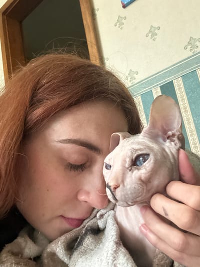 A close-up, indoor portrait shows a person with reddish-brown hair holding a hairless cat. The person's face is partially visible, with eyes closed and head tilted downwards towards the cat, which is cradled in a blanket. The cat has light pink skin and striking blue eyes. The background features a wall with striped wallpaper and a wooden doorway. The scene appears to be a personal moment of affection between the person and their pet.