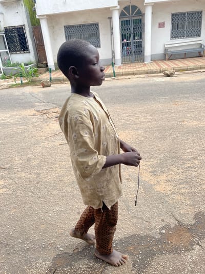 A young boy stands on a paved street, looking upwards. He wears a light-colored, patterned tunic and brown, leopard-print pants. His hands are clasped in front of him, holding a thin, dark object, possibly a twig. He is barefoot, with one foot visible in the foreground. The street is a rough, grey asphalt with some cracks and debris. In the background, a building with white walls and barred windows is visible. The building has large pillars at the entrance and decorative tiling along the sidewalk. The scene appears to be in an urban or semi-urban area. The lighting suggests it is daytime, and the weather seems to be fair. The boy's gaze upward could indicate curiosity, contemplation, or a reaction to something above him. There is no visible text in the image.