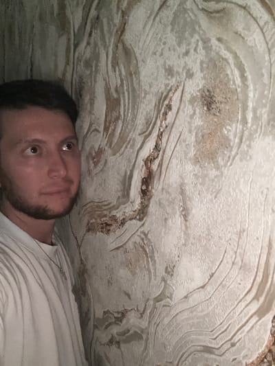 A man with dark hair and a beard is leaning against a textured wall. He is wearing a light-colored t-shirt. The wall has a rough, layered texture with swirling patterns in shades of white, beige, and brown. The man is looking upwards and to the right, with his eyes open. The scene appears to be indoors, possibly in a cave or a building with decorative wall treatment. The location is identified as Mirny, Russia.