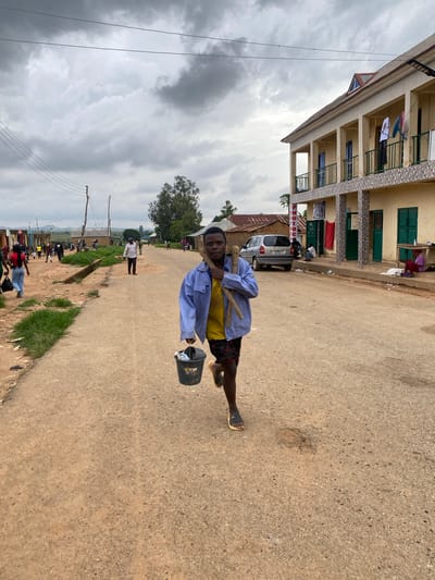 This image captures a vibrant street scene in Riyom, Nigeria, under an overcast sky. The primary subject is a young man in the foreground, positioned slightly to the right, who is smiling directly at the camera. He is dressed in a light blue long-sleeved shirt worn over a yellow garment, dark shorts, and dark sandals. He carries two wooden sticks or poles balanced on his left shoulder and holds a black bucket with some white contents in his left hand.

The setting is a wide, unpaved or lightly asphalted road that appears to be a local thoroughfare. To the left, a multi-story building with a light cream-colored facade, green window frames, and balconies is prominent. Laundry is visible hanging on several balconies, suggesting residential use. A silver car is parked in front of this building, partially obscured by a vertical red and white sign that clearly displays the text "PHOTO".

Numerous other people are scattered along the road and its grassy verges in the midground and background, engaged in various daily activities, some walking and others standing. In the background, additional buildings with corrugated iron roofs and dense trees are visible, hinting at a mix of residential and perhaps commercial activity. Wooden utility poles with power lines run along the right side of the road, receding into the distance. The cloudy sky indicates a daytime scene, though without bright sunshine. The overall impression is that of an active, everyday street environment in a local community.