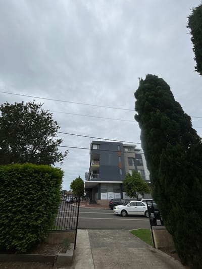 A vertical landscape features a modern apartment building with gray and beige accents on a cloudy day. Power lines stretch across the sky. Trees flank the top and bottom, and a parked white car is visible on the side street. The image is framed by a sidewalk on the right and a wrought iron fence in the foreground.
