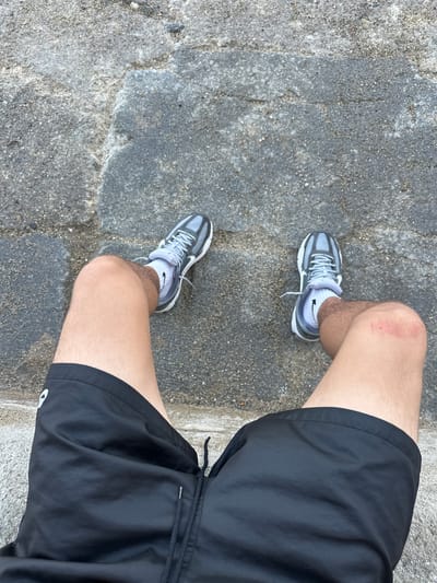 The image shows a person's legs, from the knees down, wearing black athletic shorts and grey running shoes. The person is sitting on a stone surface that appears to be part of a walkway or path. There is a visible abrasion or redness on the right knee. The environment is outdoors, with the stone ground as the primary element. The location in the city of Collioure, France is not discernible from the visual information provided in the image.