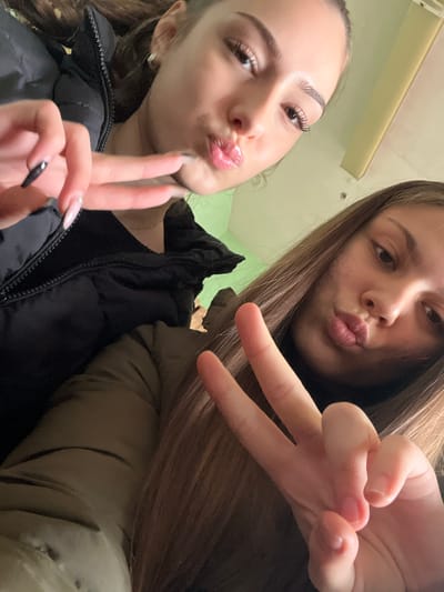 This image depicts two young females in an indoor setting, located in Cherven Bryag, Bulgaria. Both individuals are positioned for a close-up photograph. The female in the upper left wears a black puffer jacket and a black top. Her left hand, adorned with long, light-colored artificial nails (one with a black tip), is raised to display a "peace sign" gesture. Her lips are pursed. The female in the lower right wears an olive-green puffer jacket. Her right hand, displaying natural nails, is raised to form a "peace sign." Her lips are also pursed. Both subjects are looking towards the camera. The background consists of an indistinct light-colored wall, possibly light green or yellow, with a linear architectural feature or light fixture visible in the upper right portion.