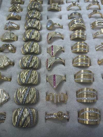 This media file is a close-up, eye-level shot of a collection of gold rings displayed on a grey fabric surface, likely in a jewelry store or personal collection. The rings are arranged in rows and columns, with some individual pieces and some pairs. They vary in design, with some featuring intricate patterns, while others are adorned with small, clear gemstones that appear to be diamonds or cubic zirconia, and a few have red stones, possibly rubies. The gold appears to be yellow gold, and the lighting highlights the shine and detail of each piece. There are no people visible in the image, nor any specific time of day, weather, or strong emotional cues. The setting is generic, focusing solely on the jewelry. There is no visible text in the image. The context provided indicates the location is Yunus Emre, Turkey, which might suggest a particular style or origin of the jewelry, but this is not evident from the visual information alone.