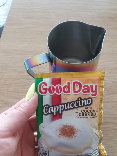 A hand with light green nail polish holds a sachet of "Good Day Cappuccino with Cocoa Granule" coffee in front of a metal frothing pitcher. The sachet features the Italian flag colors and an image of a frothy cappuccino. The background is a light brown wooden surface. The image does not provide any information that can be used to determine a location in Kheyrabad, Iran.