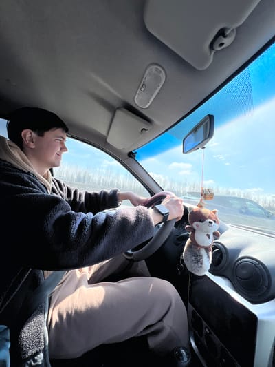 The image shows a young man sitting in the driver's seat of a car. He is holding the steering wheel and looking upwards, with a slight smile on his face. He is wearing a dark blue fuzzy jacket over a beige hooded sweatshirt. The car's interior is visible, including the dashboard, sun visors, and rearview mirror. Hanging from the rearview mirror is a small plush toy of a dog connected with a string to a small cube. The car appears to be parked, with a view of trees and a bright sky visible through the windshield. There are no identifying features that would place this image in the city of Russkie Kirmeni, Russia.