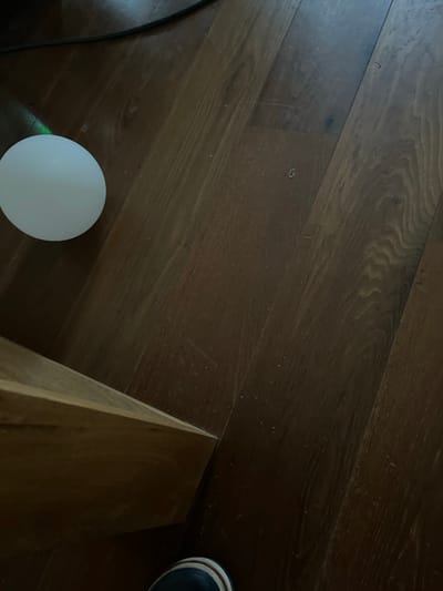 The image depicts a close-up, overhead view of a dark wooden floor, possibly oak, with visible wood grain patterns. In the upper left quadrant, a translucent white spherical object is positioned on the floor. A faint, iridescent light, possibly from a light source or a reflection, emanates from the left side of this sphere, casting a subtle green hue. To the left of the sphere, a black cable is visible, lying on the floor and extending upwards and out of the frame. In the lower left corner, the edge of a light-colored wooden furniture piece is present, angled diagonally into the frame. In the very bottom center of the image, a portion of a person's foot is visible, wearing a dark shoe with a white and red striped band. There are no indications of the location being in the city of Orekhovka, Russia, or any other specific geographical context. The scene appears to be indoors, in a domestic or office setting, with no activity or interaction occurring. The overall lighting is dim, with the primary light source seemingly originating from the left.
