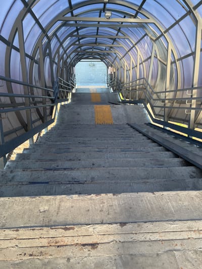 The image shows a covered pedestrian overpass with a blue translucent roof and a metal frame. A set of concrete stairs leads down from the overpass, with metal handrails on both sides. Yellow tactile paving is visible on some of the stairs. The overpass is located in Kalynivka, Ukraine, and appears to be part of the city's infrastructure. The scene is captured from a downward-looking perspective, focusing on the stairs and the entrance to the overpass. The lighting suggests it is daytime, with shadows cast by the structure. There are no people or animals visible in the image. The environment appears to be urban, with glimpses of a street or pathway at the bottom of the stairs.
