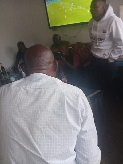 A gathering of people is watching television. A bald man in a light blue patterned shirt is in the foreground, his back to the viewer. Another man wearing a white sweatshirt is looking at the television screen. The television screen shows a green background and appears to have graphics resembling a sporting event. Water bottles and other items are on the floor.