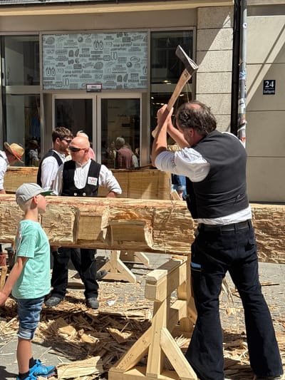 The image depicts an outdoor scene in Munich, Germany, specifically on Kaufingerstraße, as indicated by the building number "24" visible on a street sign. The central activity involves a man vigorously swinging an axe at a large wooden beam, which is supported by wooden saw horses. He is wearing a dark vest over a white shirt and dark corduroy pants. To the left, a young boy in a light blue t-shirt and denim shorts watches the action, wearing a grey baseball cap and blue athletic shoes. Several other individuals are present in the background, some wearing traditional Bavarian attire, suggesting a cultural or historical event. The ground around the wooden beam is strewn with wood shavings, indicating recent work. Behind the participants, the facade of a commercial building with large glass windows is visible, displaying merchandise and signage, including "Urban Outfitters" and "BMZ". The bright sunlight and clear skies suggest a daytime setting.