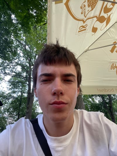 A young, light-skinned male with brown hair and light brown eyes is pictured in a close-up, front-facing selfie. He is wearing a white t-shirt and has a black strap across his left shoulder. The background consists of green trees and a white umbrella with orange lettering. The scene appears to be outdoors, possibly in Jihlava, Czechia, given the umbrella's design which resembles those commonly found at outdoor seating areas of establishments like beer gardens or cafes. The lighting suggests it is daytime.