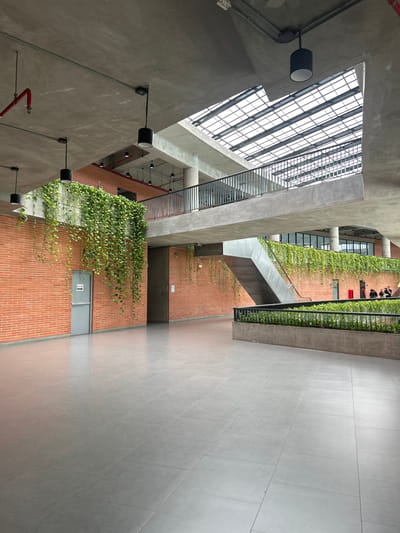 An interior view captures the modern architecture of a large building. The scene is dominated by exposed concrete ceilings and walls, accented with exposed brickwork. Natural light streams in from a large, grid-patterned skylight at the top of the image. Suspended black cylindrical lights hang from the ceiling. Lush green vines cascade down from elevated walkways and planters, adding a biophilic design element.
In the foreground, a wide expanse of grey tiled flooring stretches across the image. A grey door with a "PUSH" sign and an arrow pointing left is visible on the brick wall to the left. Further into the space, a multi-level structure is evident, with a staircase leading upwards. A black metal railing lines the edge of an upper walkway.
In the middle ground, a few individuals are present, appearing to be standing and conversing near a red rectangular object. The overall impression is of a spacious, contemporary public building with an emphasis on natural light and greenery. The location is identified as a building within Tha Khlong Town Municipality, Thailand.