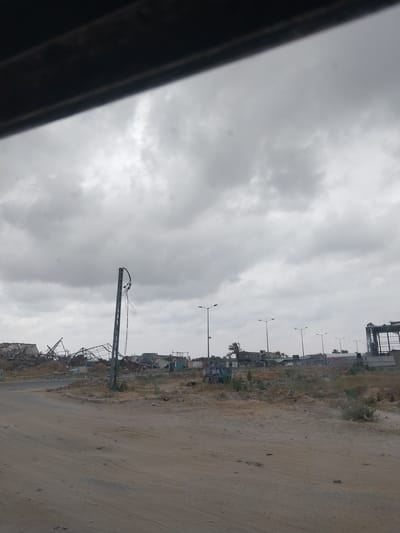 The image shows a street scene in al-Qarara, Palestinian Territories, under an overcast sky. Along the side of the street, there is rubble and debris from demolished buildings. Utility poles stand among the destroyed structures. The road itself is dusty and worn. A dark object is visible in the extreme left of the frame, partially obscuring the view. There are no people visible.