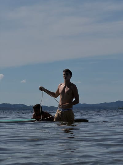 The image shows two men on a body of water, possibly a lake or sea, against a backdrop of a mountain range and sky. One man is standing shirtless, holding a rope and facing forward, suggesting he is water skiing or wakeboarding. The other man is partially submerged in the water, holding onto a board. The water shows ripple patterns. The location is most likely in General Luna, Philippines.