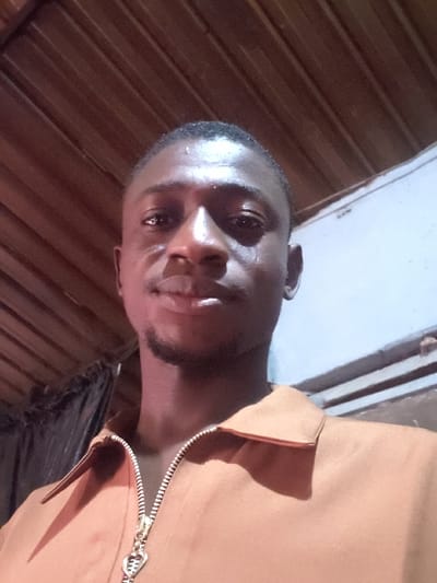 A dark-skinned man with closely cropped hair poses for a selfie indoors. The camera angle is low, capturing his face and the upper part of his brown collared shirt with a visible zipper. The lighting is warm, casting shadows on the left side of his face. A wooden wall is visible in the background, contrasting with a brighter, lighter colored wall or surface above it.