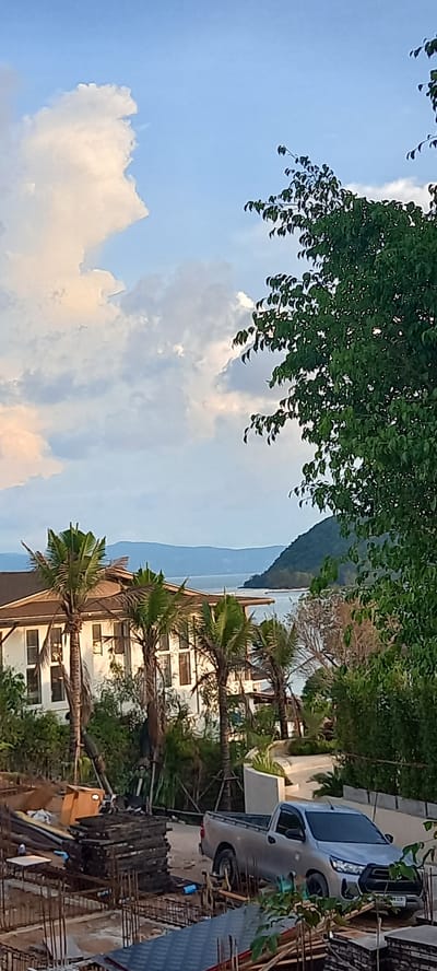 Baan Wok Tum, Thailand. A daytime, outdoor scene capturing a partial view of a building adorned with palm trees, observed from an elevated perspective, likely a construction site. To the right, a light-colored pickup truck parked adjacent to building materials. In the background, a mountain and sea visible. A mix of clouds and sky are visible to the left, partially obscured by foliage.