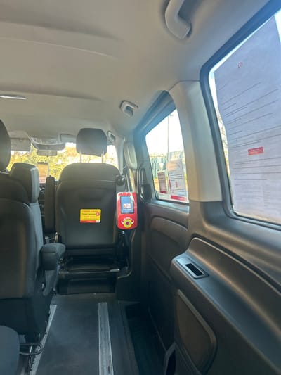 The image depicts the interior of an unoccupied commercial passenger vehicle, likely a shuttle or accessible van, positioned in Lingolsheim, France, evidenced by the visible "CTS" (Compagnie des Transports Strasbourgeois) branding on multiple elements. The perspective is from the rear passenger area, looking forward and towards the right side of the vehicle.

The interior features dark-colored, upholstered seats; the backrest and headrest of a forward-facing middle-row seat are prominently centered. Attached to the back of this seat is a red CTS ticket validation device with a circular yellow "VALIDEZ ICI" button and text indicating "TICKET UNIQUE." Directly above it, a yellow informational sticker displays "VENTE À BORD," "TICKET UNIQUE," and "TARIF UNIQUE," along with a QR code and the CTS logo. The floor of the vehicle is dark and includes visible metal tracks, suggesting adaptable seating arrangements. The right-hand side panel is dark-colored with a recessed handle and a speaker grille.

Several white informational papers, some bearing the CTS logo, are taped to the right-side windows. Through the windows, bright sunlight illuminates an exterior scene showing deciduous trees with bare branches, some green foliage, and distant urban structures, indicating a daytime outdoor setting. No individuals are present in the vehicle, and no specific actions or interactions are depicted.
